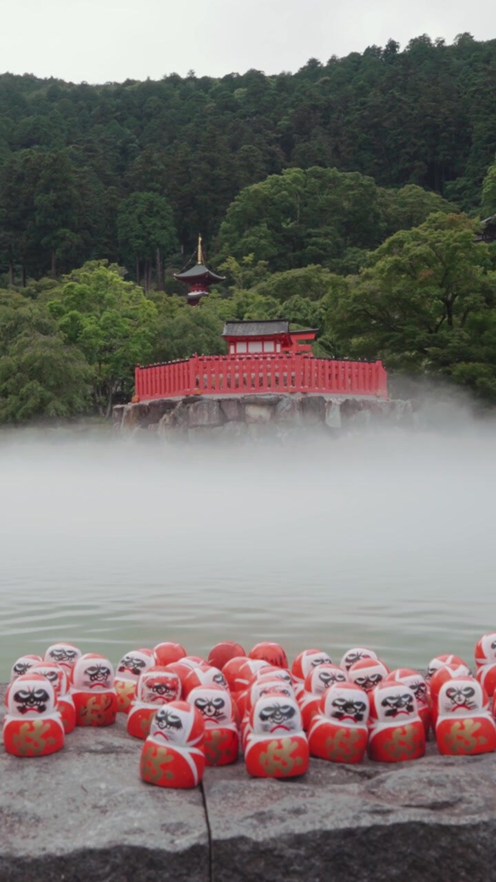 Video con orientamento verticale di bambole Daruma al Tempio Katsuoji, con un caratteristico tempio buddista rosso e una pagoda circondati da una fitta foresta verde e nebbia suggestiva