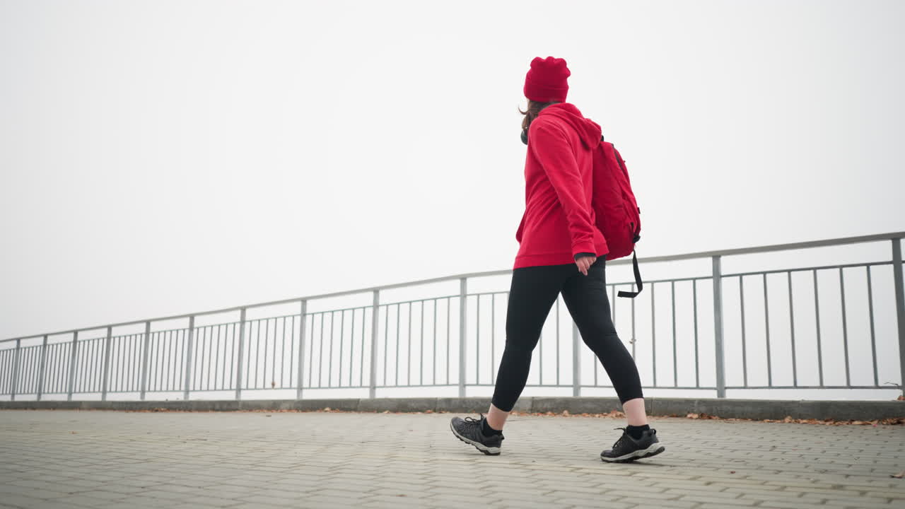 Side view of freelancer in red sweater and head warmer, walking confidently along interlocked pathway carrying bag over shoulder, surrounded by serene foggy atmosphere and autumn leaves