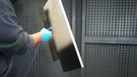 man applying black anti-corrosion powder coating to metal sheet using a spray gun in factory