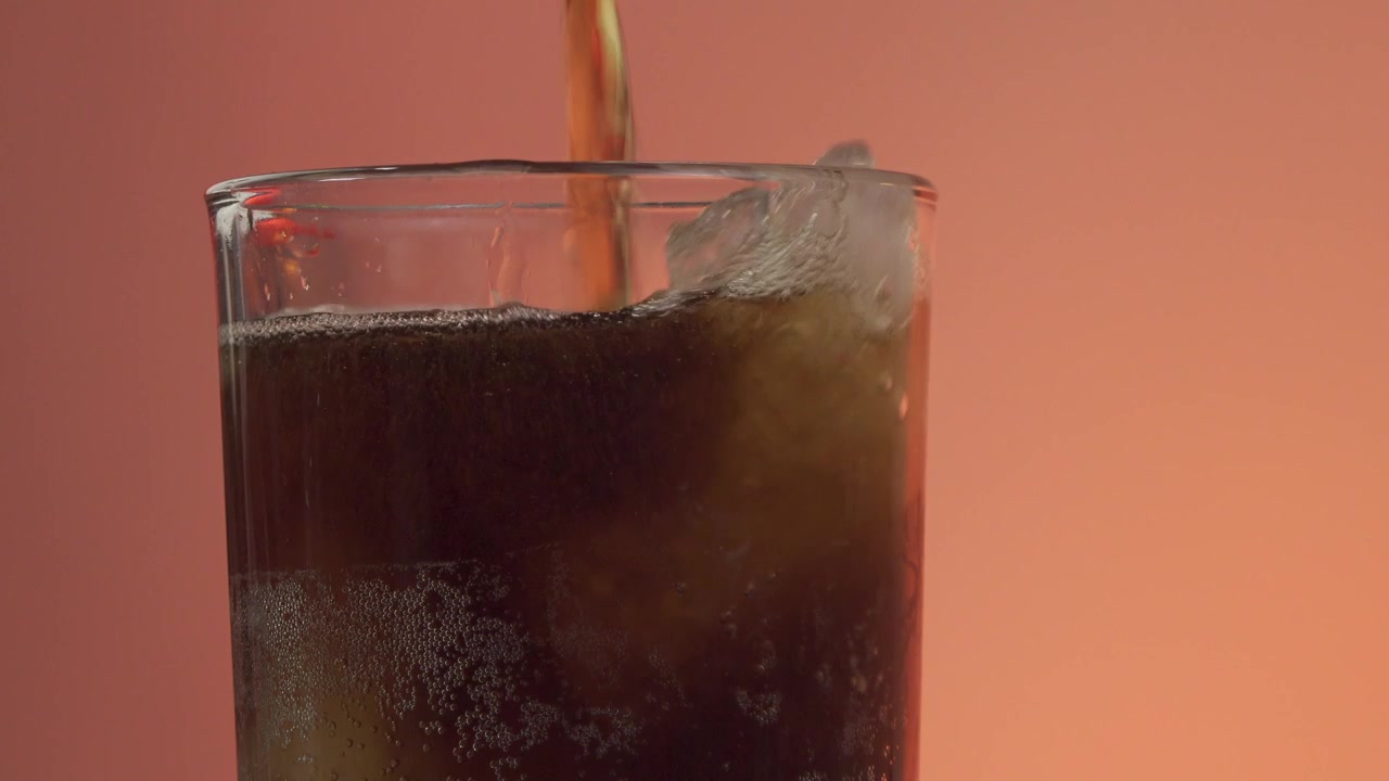 Versamento di soda in un bicchiere con ghiaccio su uno sfondo arancione vivace.