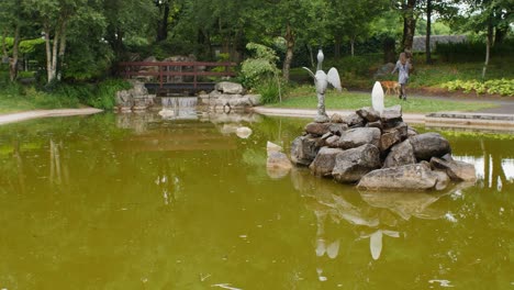 Modelo-Femenino-Camina-Con-Perro-Por-Un-Estanque-Sereno,-Cruzando-Un-Puente-De-Madera-Cerca-De-Una-Pintoresca-Cascada