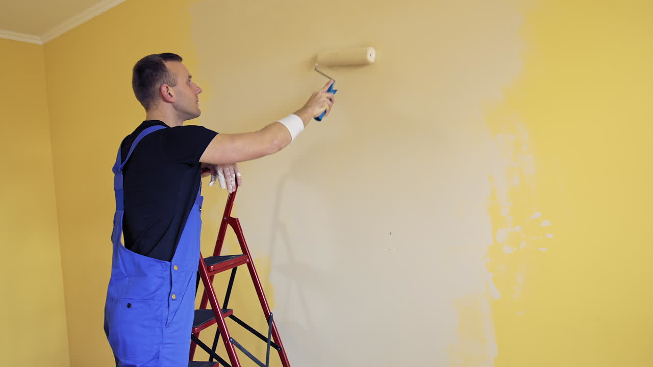 Man in blue overalls paints room. Worker paints the walls in the room with a paint roller on a ladder. Room decoration process into new color.