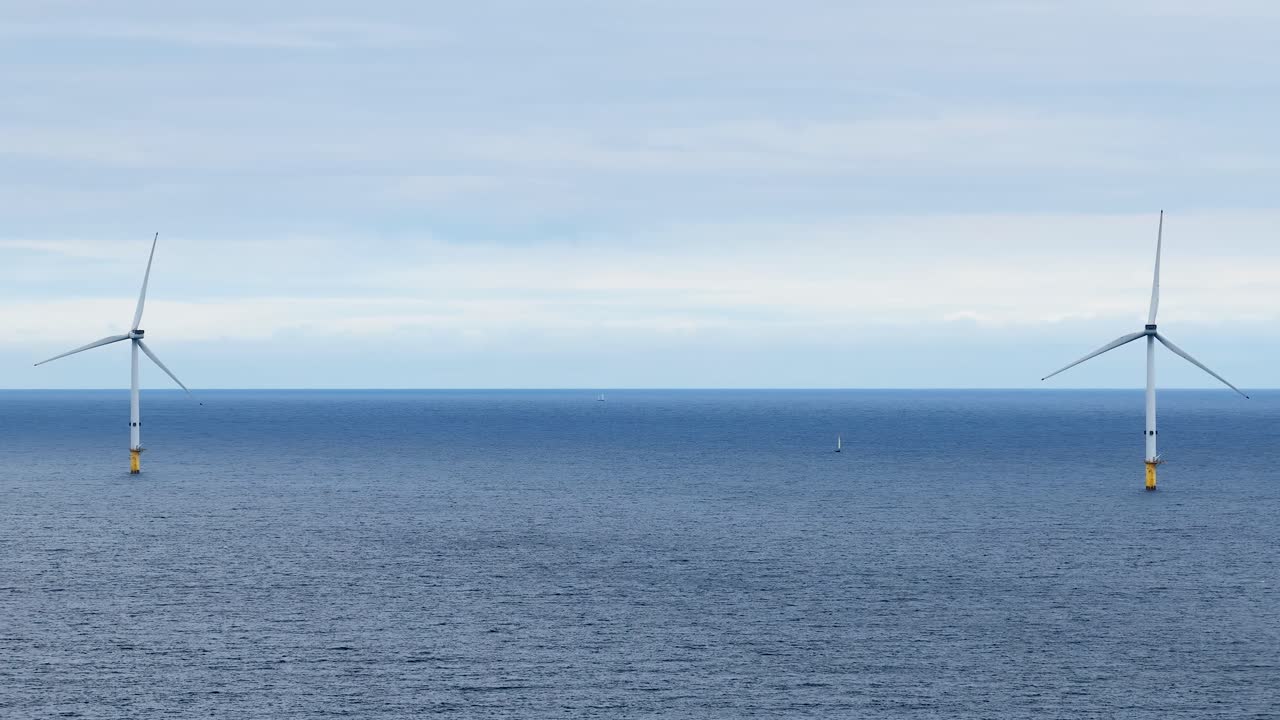 Two offshore wind turbines rotate steadily in the open sea under overcast daylight, captured with a static wide shot and minimal camera movement