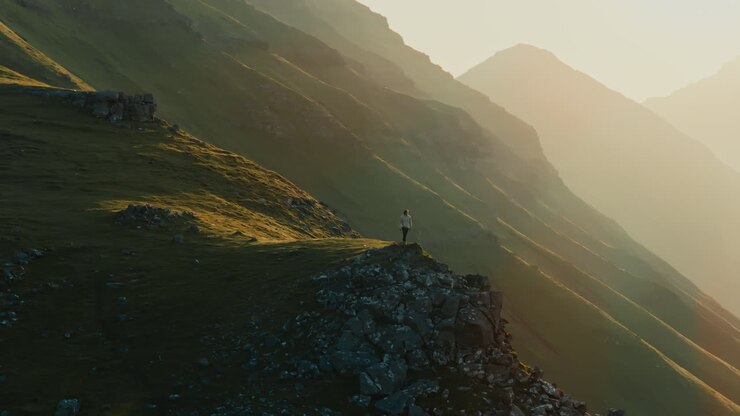 Person standing on a rocky peak overlooking a vast green mountain landscape during golden hour