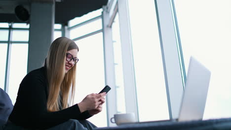 young female freelancer works near window. beautiful woman sitting in front of a laptop using a mobile phone to type messages. profile view.