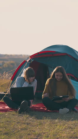 positive women hikers with tablet and laptop work sitting on red plaid by tent and bonfire on river bank in autumn evening