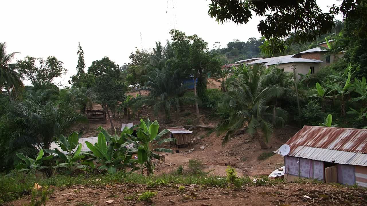 Landlig bebyggelse på en bakketop i det centrale Ghana med små huse og bliktag omgivet af frodig tropisk vegetation, palme- og banantræer samt jordstier i et roligt landsbylandskab