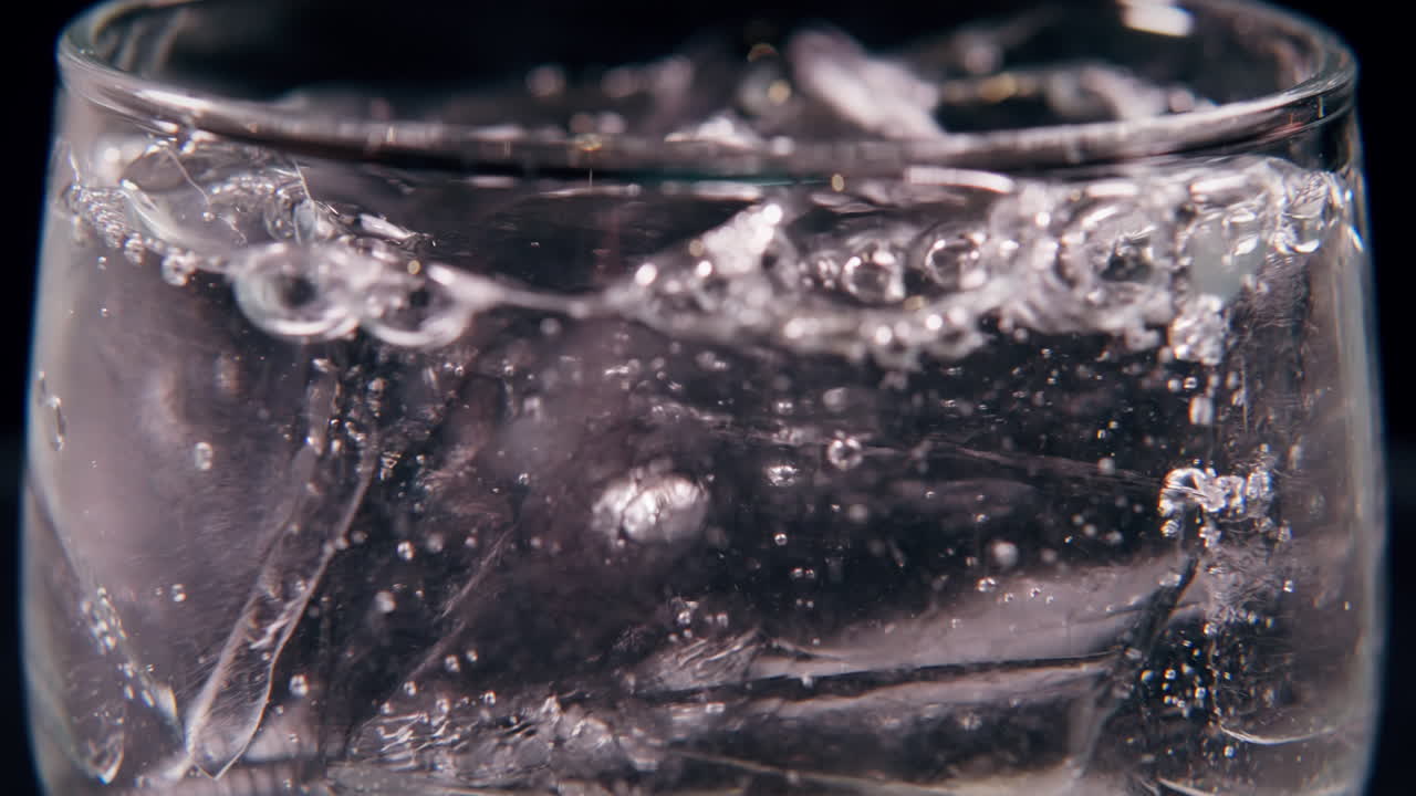 Scatto macro di acqua versata su cubetti di ghiaccio in un bicchiere trasparente, catturato in slow motion cinematografico con illuminazione drammatica.
