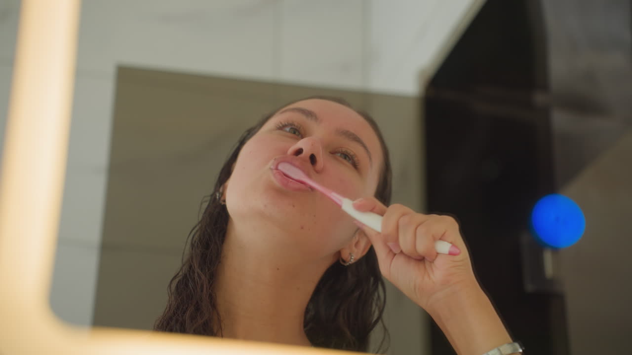 Playful female cleans teeth images. Evening scene featuring woman and glowing mirror reflections. Modern room illuminated by soft light showing woman happily brushing her teeth during nighttime