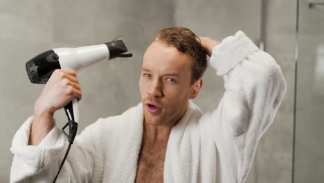 handsome man in a white bathrobe dries his hair with a hair dryer