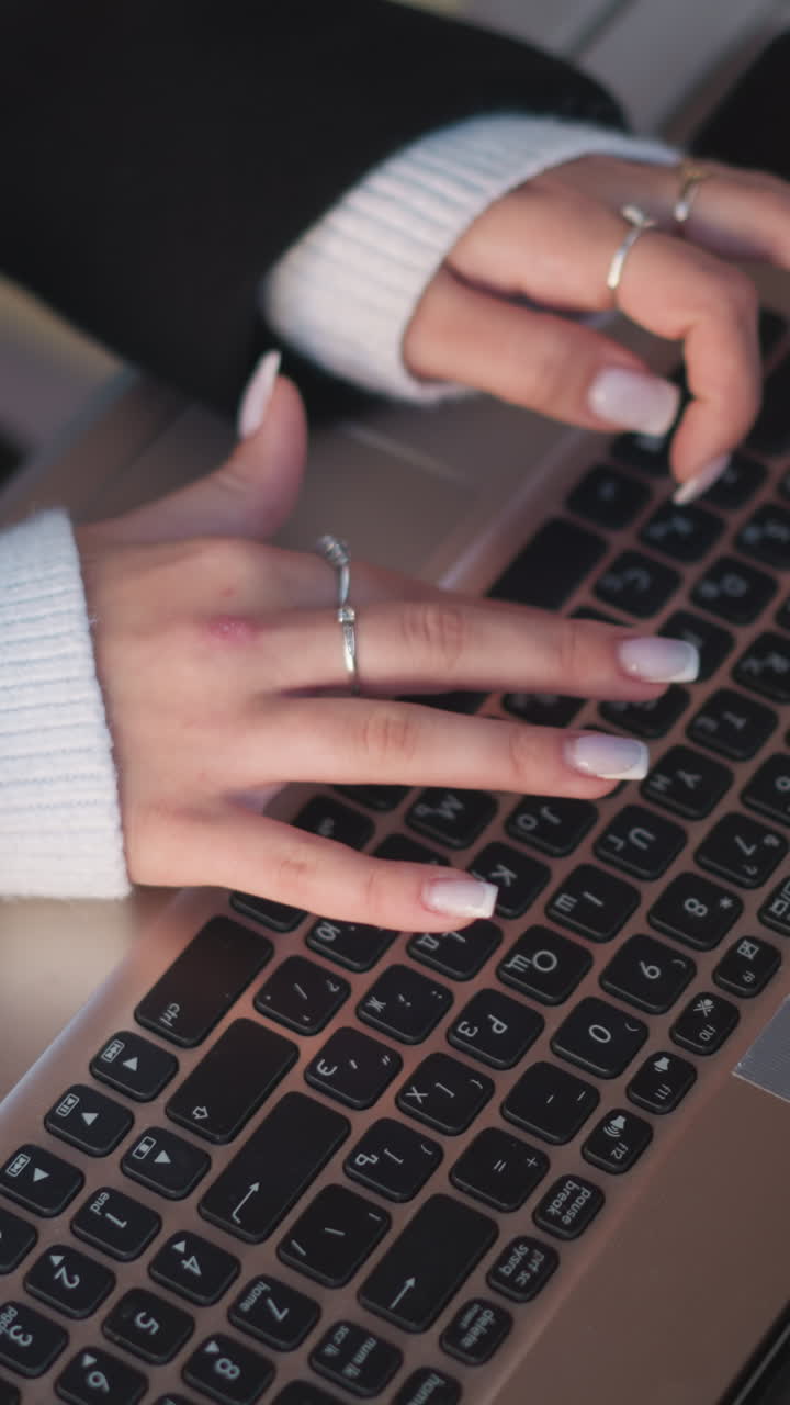 primer plano de manos con uñas pulidas escribiendo en el teclado de la computadora portátil, adornado con anillos de plata y cómodas mangas de suéter, la computadora portable se coloca en una elegante mesa de madera en un espacio de trabajo al aire libre