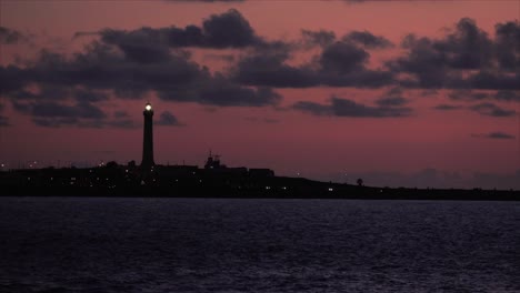 Faro-Recortado-Contra-Una-Puesta-De-Sol-Roja-Ardiente,-Un-Suave-Resplandor-Sobre-El-Sereno-Paisaje-Marino-Y-El-Horizonte