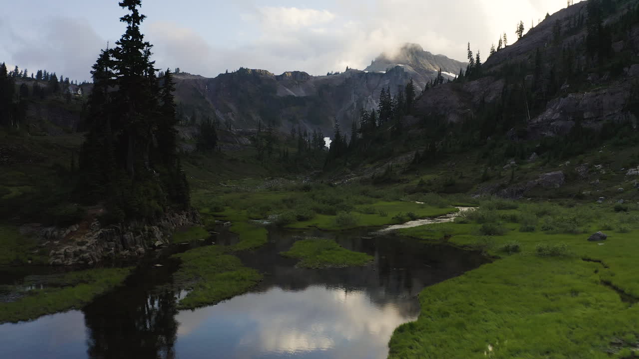 Rocky PNW alpine landscape reflects in shimmering meadow lake with lush green forest and grass, aerial dolly