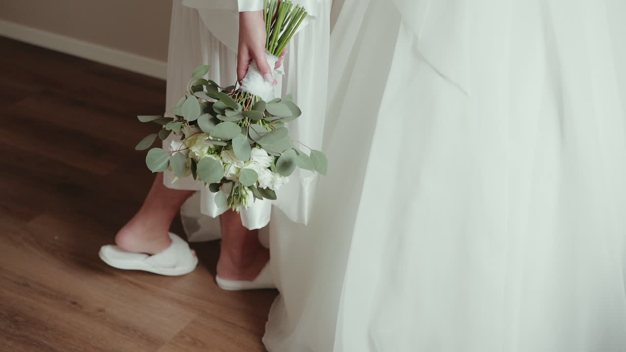 bride holding bouquet while walking in slippers next to wedding dress on wooden floor