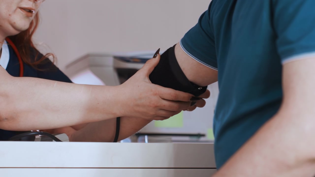 Infermiera bianca che applica il bracciale per la pressione sanguigna sul braccio del paziente, con un primo piano delle mani e del bracciale nero, in un ambiente clinico sereno, tecnica precisa, paziente maschio bianco seduto alla scrivania, attenzione sul dispositivo e sui valori.