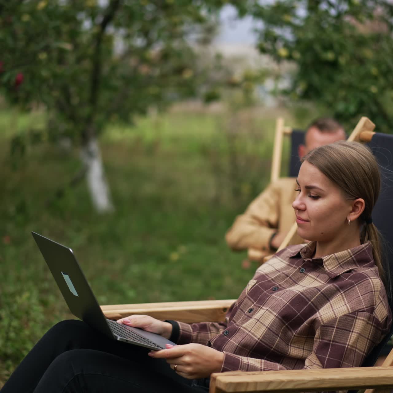 Busy freelancing female employee typing on her laptop. Lady finishes work leaning backwards in the chair. Man using smartphone at backdrop