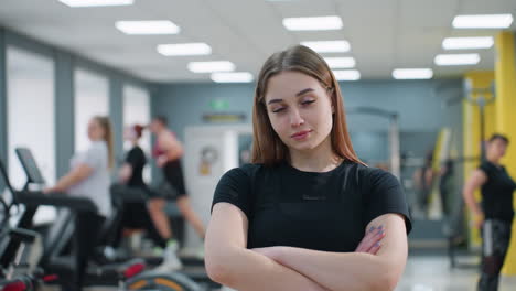 confident young woman standing with arms crossed inside bright modern gym, posing calmly while other individuals exercise on treadmills and strength equipment in background under white ceiling lights