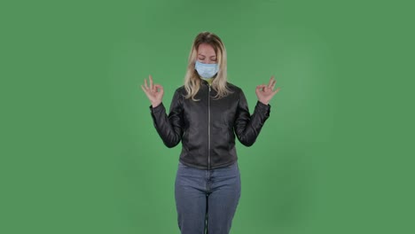 의료 보호 얼굴 마스크를 입은 아름다운 젊은 여성의 초상화는 카메라를보고 휴식을 취하고 명상하고 있습니다. 검은 재과 청바지를 입은 느슨한 머리카락을 가진 금발은 스튜디오에서 녹색 화면에 있습니다. 건강 보호 코로나 바이러스 개념