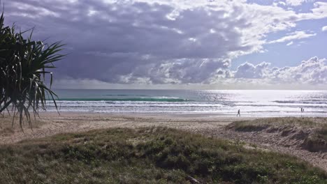 Menschen-Gehen-Am-Wunderschönen-Strand-Entlang,-Während-Glitzernde-Blaue-Wellen-Auf-Dem-Sand-Brechen