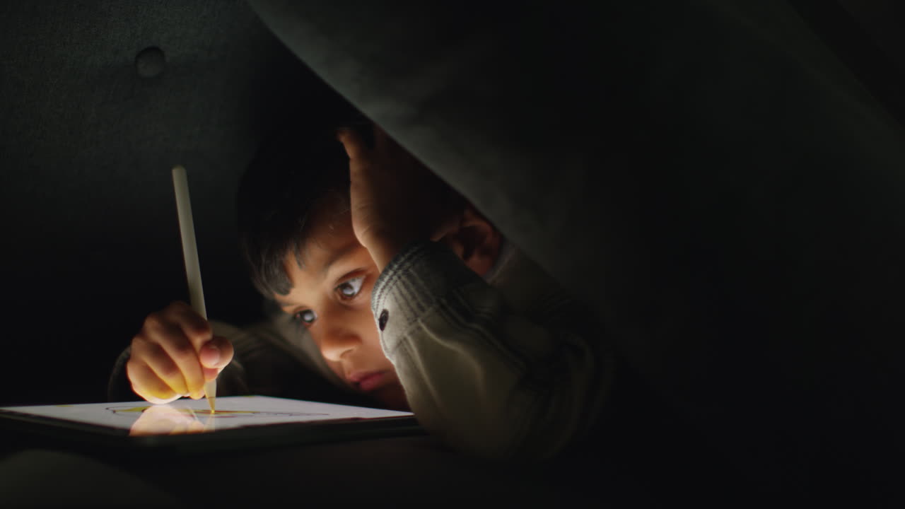 Young Boy In Home Made Camp Made From Cushions Playing And Drawing With Digital Tablet And Stylus At Night 4