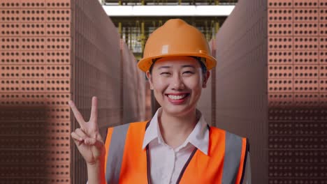 close up of asian female engineer with safety helmet smiling and showing peace gesture while standing with red brick packed in stacks are stored