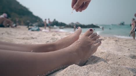 man-sprinkles-pinch-of-sand-on-woman-legs-lying-on-beach