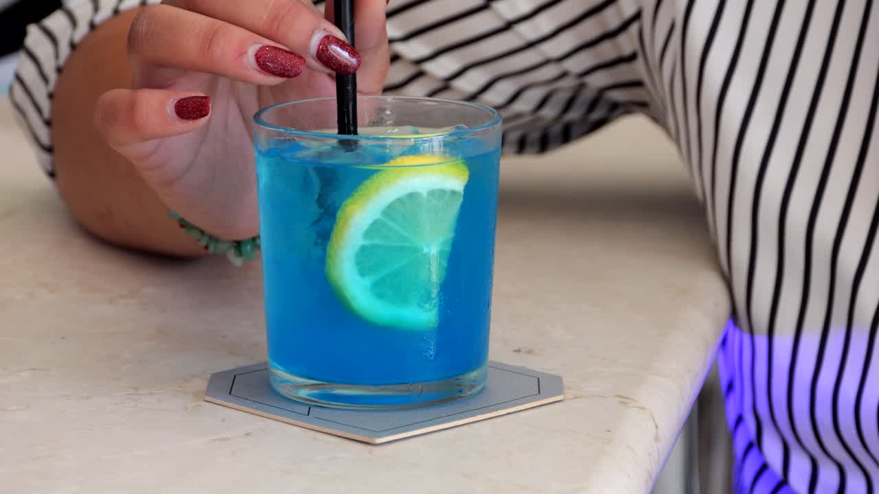Blue cocktail with lemon slice being stirred by woman's hand, black straw highlighting vibrant bar setting and refreshing beverage details