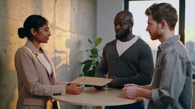 Extending woman in beige blazer guiding manila folder to two colleagues at office bay for handover
