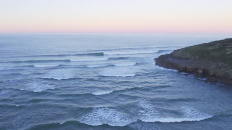 Gentle-waves-along-rugged-coast-at-sunset-create-picturesque-scene-AERIAL-4K