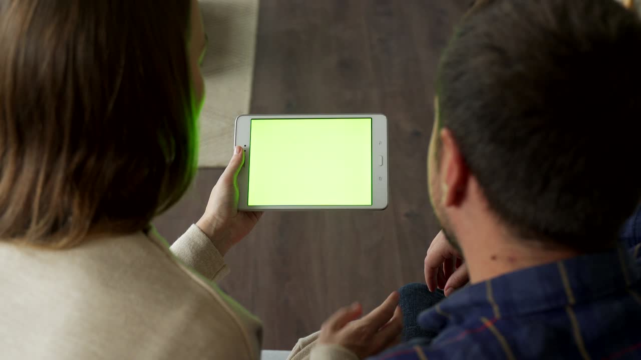 una pareja joven sentada en un sofá se puede ver sobre su hombro usando un diseño de pantalla de tableta verde. el concepto de pantalla verde