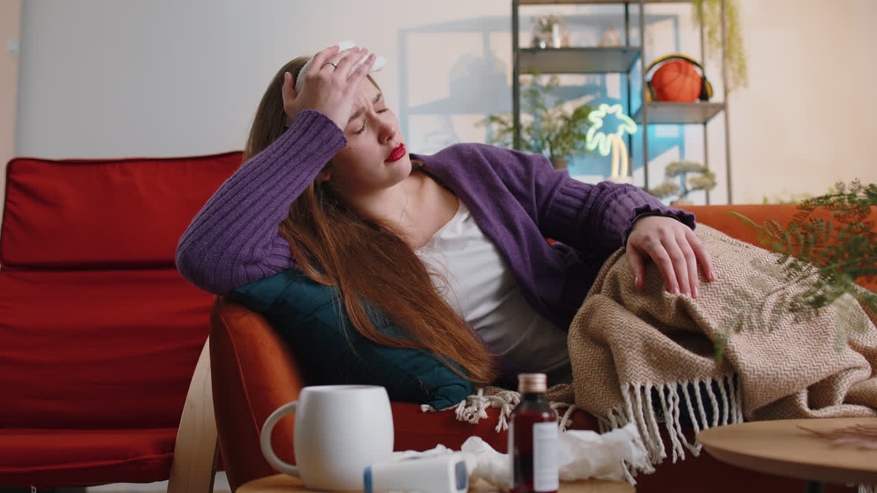 Sick ill young woman suffering from cold allergy lying on home sofa sneezes wipes snot into napkin