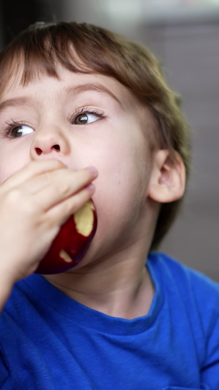 Little boy holding apple close to camera. Kid takes a bite from apples in both hands. Close up. Blurred backdrop. Vertical video