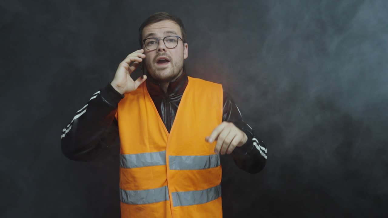 A worker wearing a reflective safety vest calls the fire department during an evacuation. Smoke and alarm lights indicate a fire emergency in an industrial area