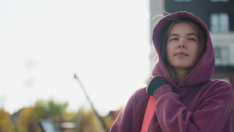 urban woman hanging pink bag on shoulders adjusting hoodie while strolling sunny street with modern building backdrop capturing fashionable confidence relaxed motion among cityscape