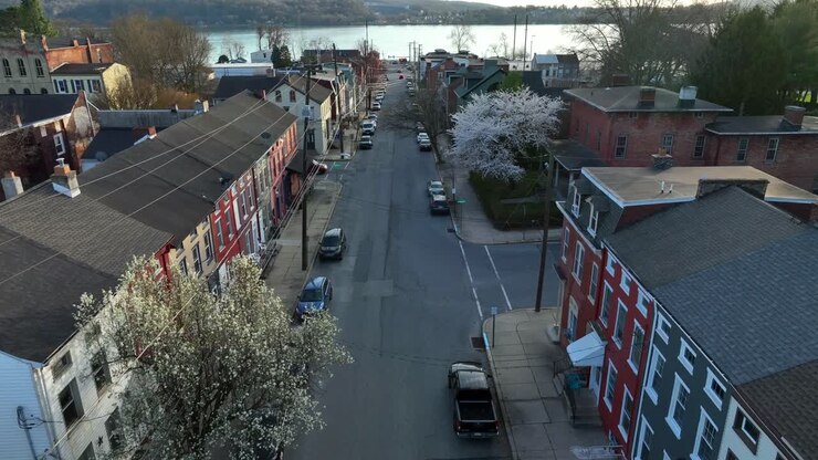 Ripresa con drone in avanzamento di una strada principale in una piccola città americana al tramonto, con i riflessi delle case e degli edifici, che termina su un lungofiume e colline oltre il fiume.