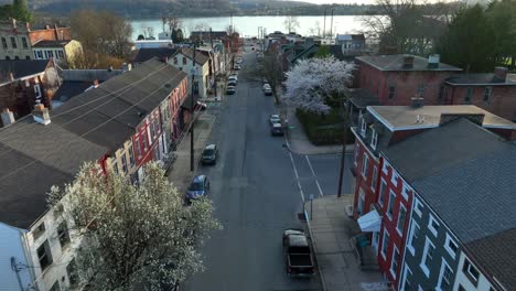 Drone-view-moving-forward-of-a-main-street-in-small-town-America-at-sunset-with-reflections-off-of-the-homes-and-buildings-ending-at-a-riverfront-and-hills-across-the-river