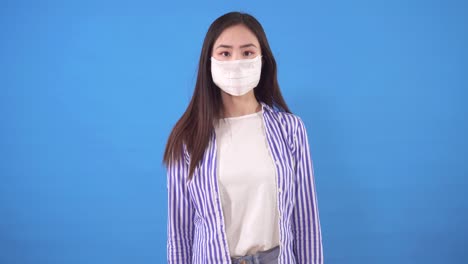 beautiful young asian woman puts a protective medical mask on her face standing on a blue background and looks at the camera