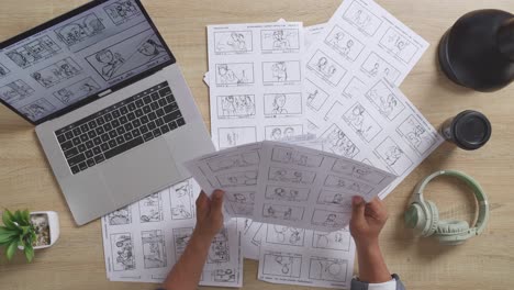 close up top view of male artist's hands holding sketching papers choosing the best storyboard for his project on the table with a laptop and headphones in the studio