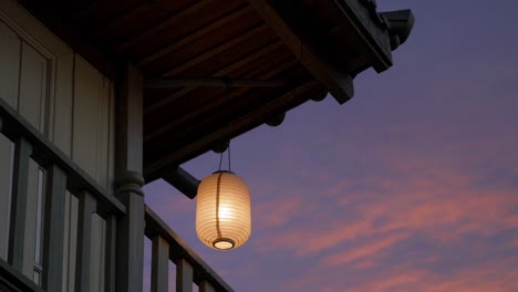 serene glow emanates from a traditional japanese paper lantern hanging from the eaves of a wooden porch, casting a warm light against the backdrop of a vibrant sunset sky