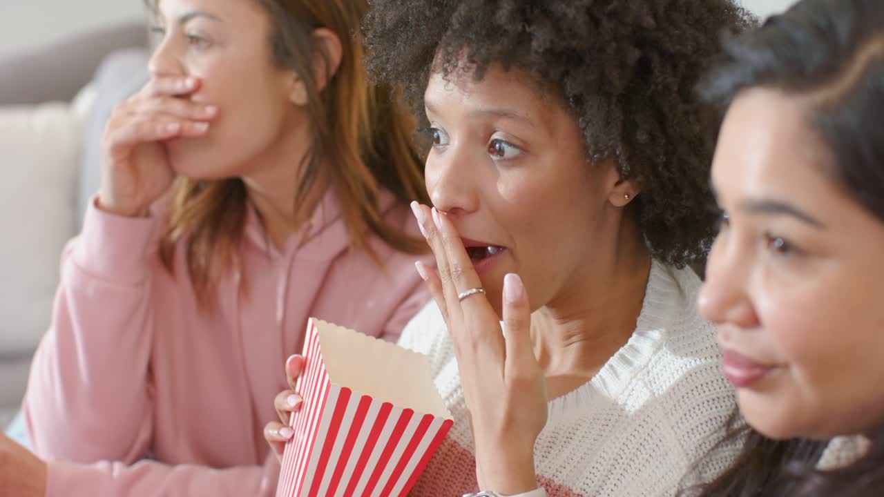 Diverse female friends snacking from popcorn boxes, reacting with gasps to on-screen twist at home