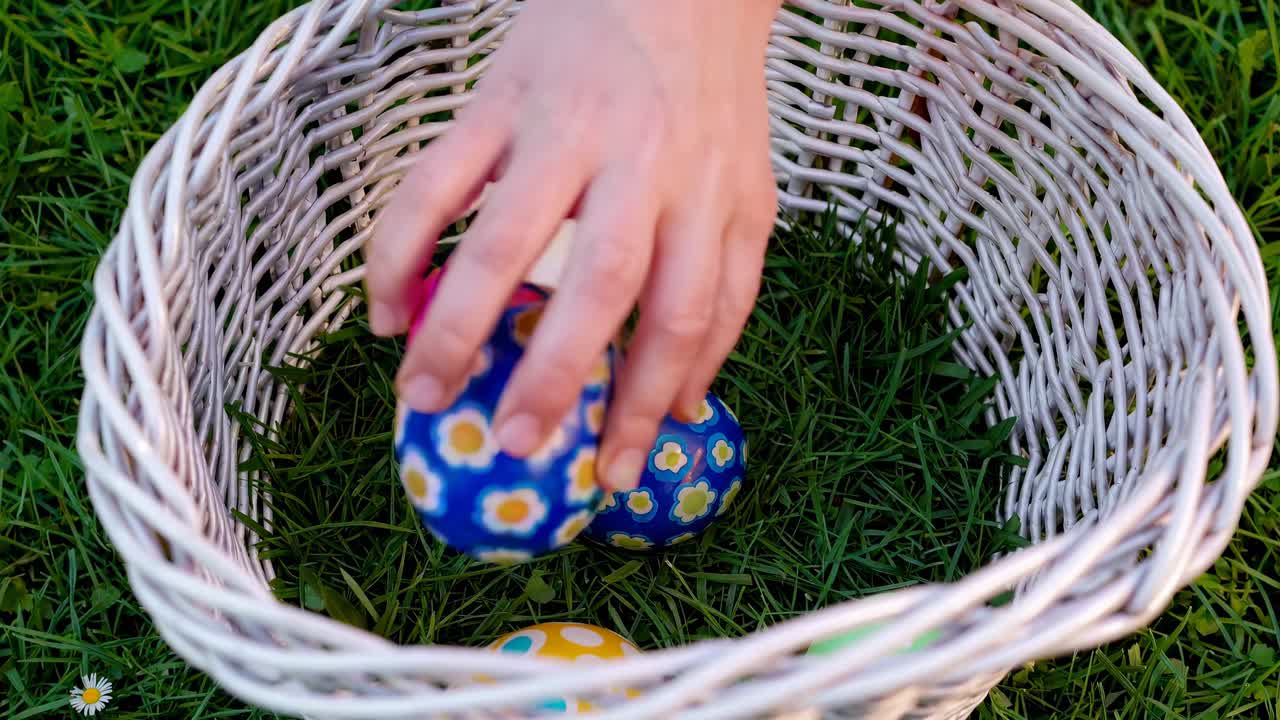 Top-down video shot of a hand placing colorful Easter eggs in a white wicker basket on grass