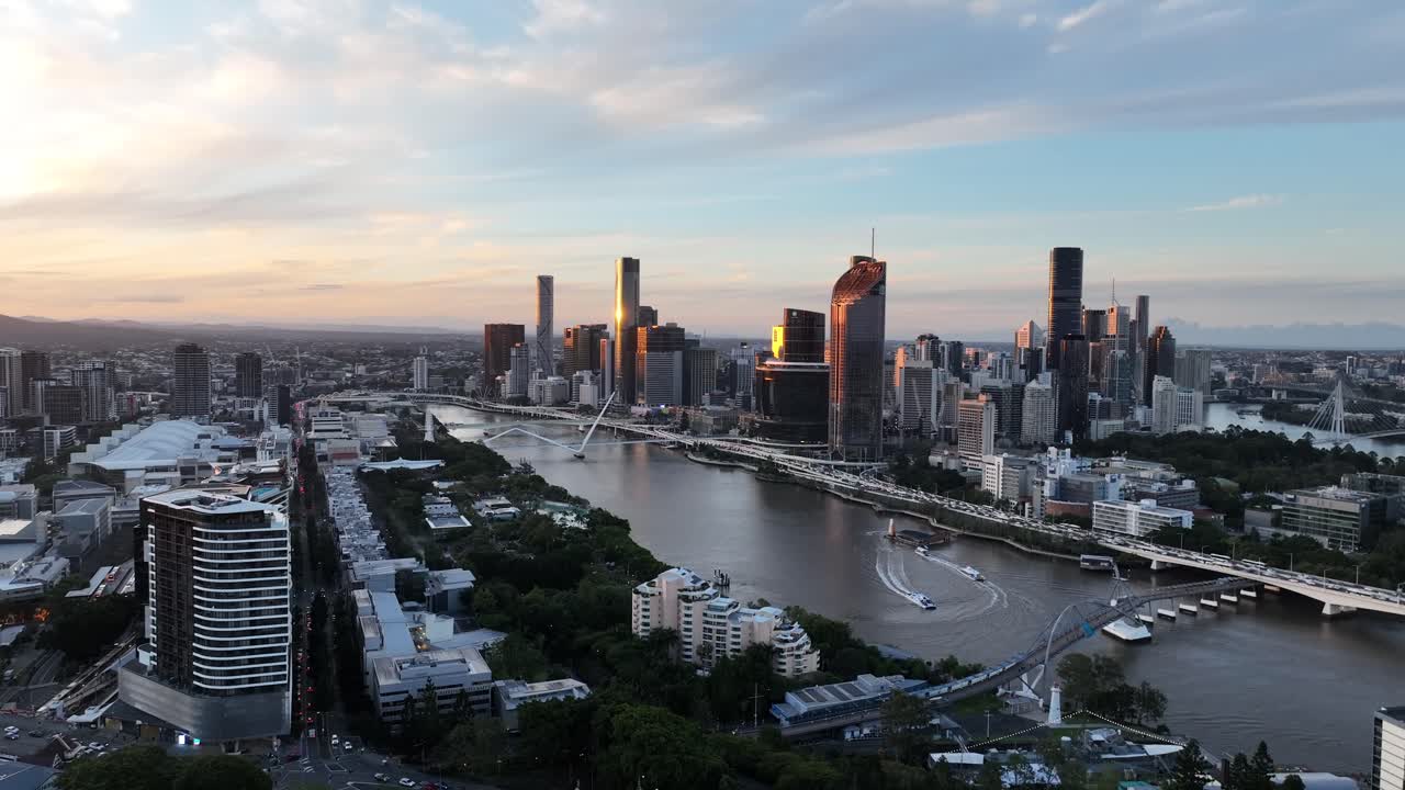 Langsom dronetracking i den gyldne time langs Brisbane-floden, der fanger CityCat-kølvandet og South Bank, mens Queens Wharf og CBD's skyline gløder i aftenlyset, Brisbane, QLD, Australien