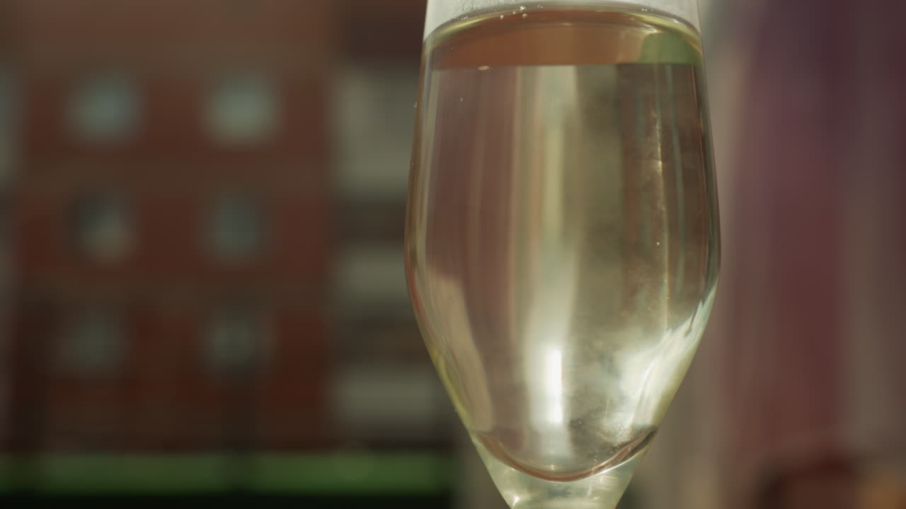 primo piano di un flûte di champagne su un balcone, con un liquido dorato e delicato che si deposita con leggere bollicine, sfondo sfocato di un edificio in mattoni, luce cinematografica calda, profondità di campo ridotta, movimento lento della camera, atmosfera romantica e accogliente