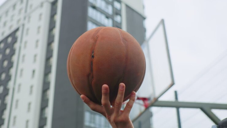 basquete girando na mão perto da cesta em área urbana, close-up da bola de couro desgastado e dos dedos se equilibrando, prédio e tabela de metal no plano de fundo, treino focado mostrando controle de movimento e coragem