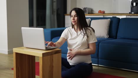 Una-Mujer-Embarazada-Vestida-De-Casa-Está-Sentada-En-El-Suelo-Y-Usando-Una-Computadora-Portátil