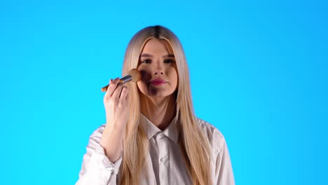 Caucasian-woman-catches-cosmetic-brush-and-puts-on-makeup-blue-studio-background