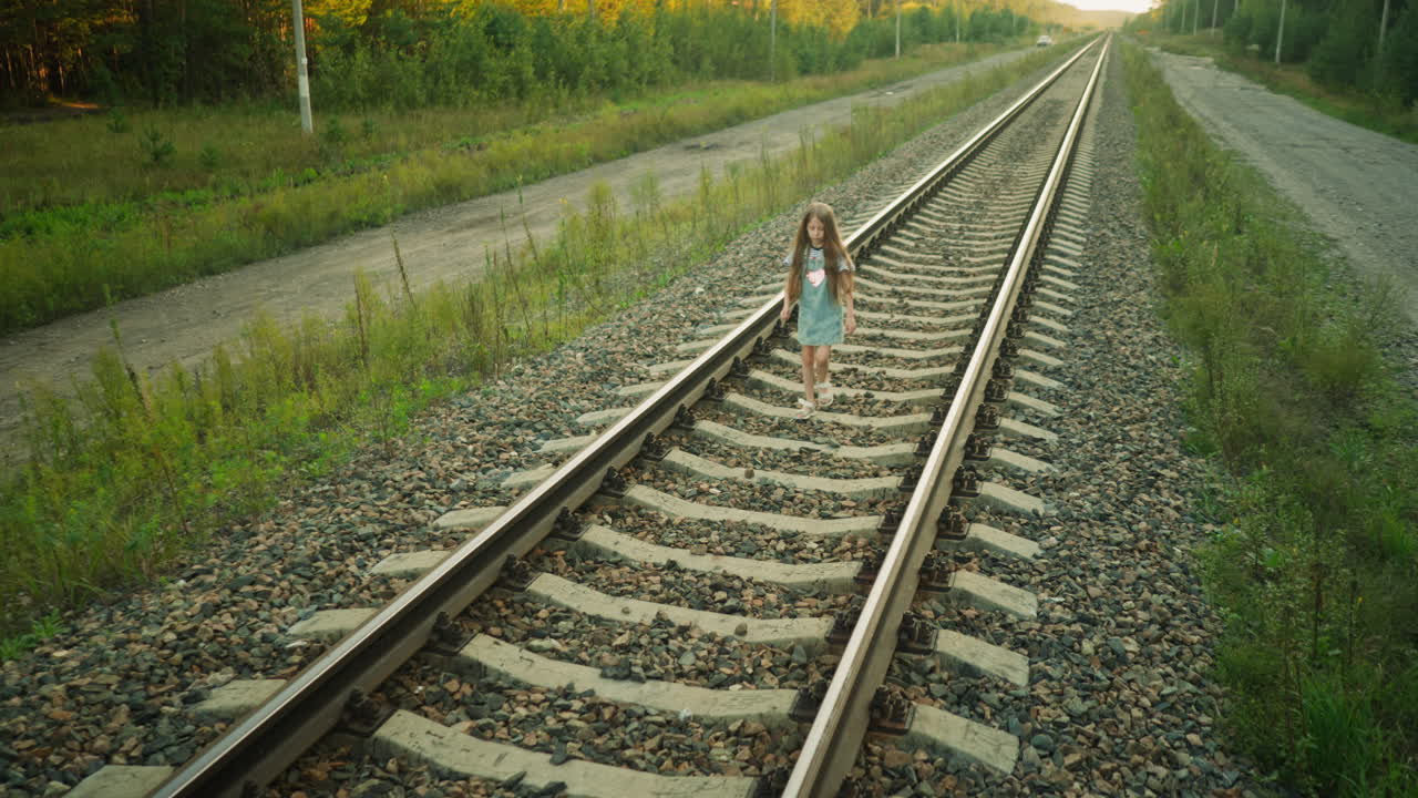 Young kid walking on railway track with hand slightly raised, dressed in denim dress and striped shirt, surrounded by rocky path, wild grass, and countryside road