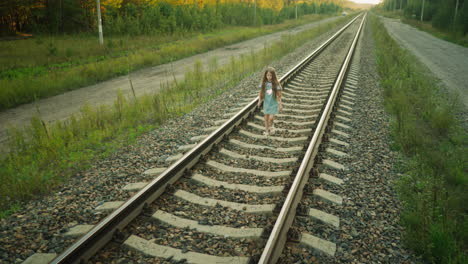 young kid walking on railway track with hand slightly raised, dressed in denim dress and striped shirt, surrounded by rocky path, wild grass, and countryside road