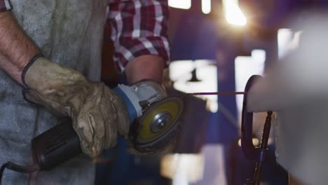 midsection of caucasian male knife maker in workshop using angle grinder