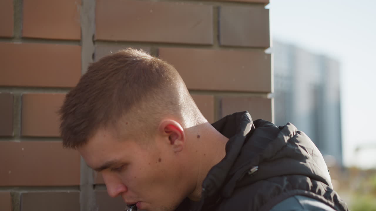 boy leaning close to brick wall secretly inhaling shisha and beginning to cough with blur view of distant office building in background under natural daylight wearing black vest and dark shirt